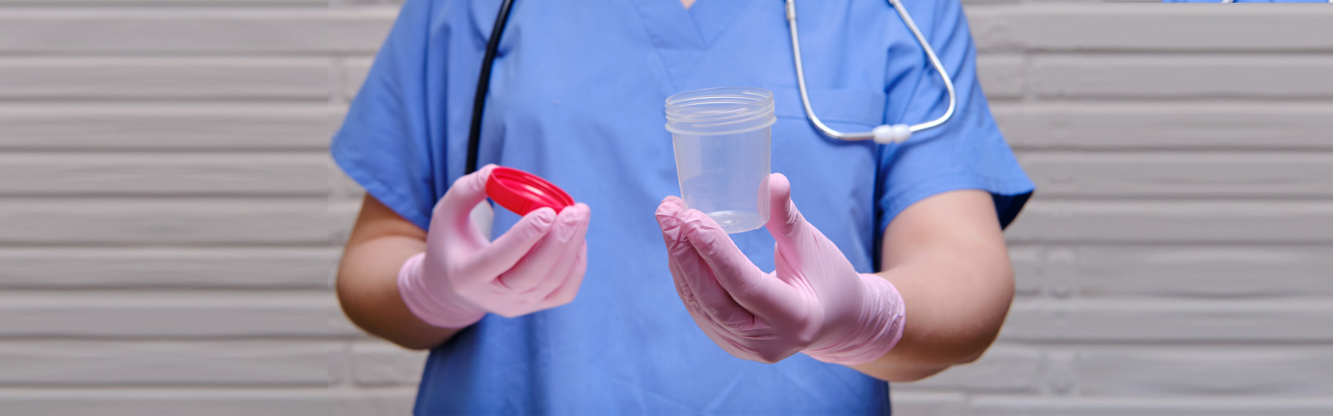 Healthcare professional in blue scrubs and pink gloves holding an open specimen container with a red lid.