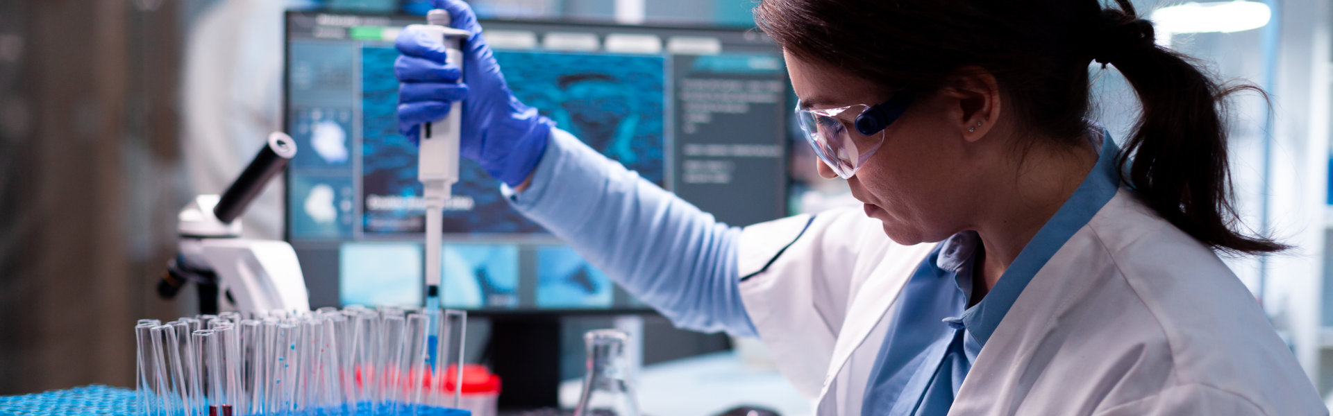 Scientist wearing blue gloves using a pipette to transfer liquid into test tubes in a laboratory with microscope and monitor screens.